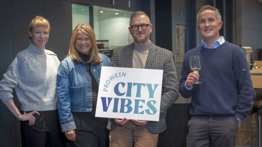 Vier Personen stehen zusammen und halten ein Schild mit der Aufschrift 'ProWein City Vibes'.