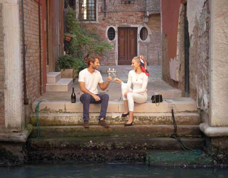Zwei Personen stoßen mit Gläsern an, während sie auf einer Treppe am Wasser sitzen.