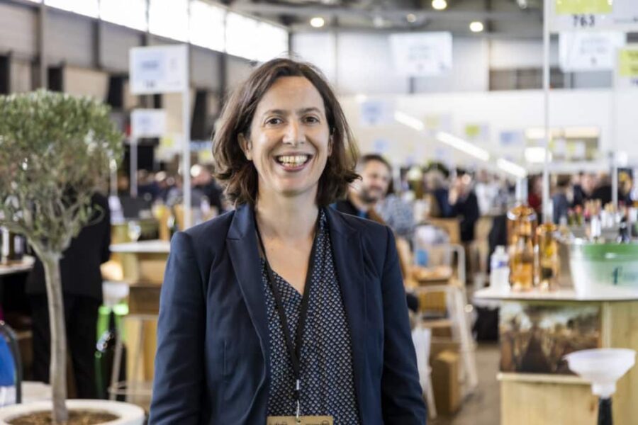 Frau mit braunen Haaren und Blazer lächelt in einer Messehalle mit Ständen im Hintergrund.