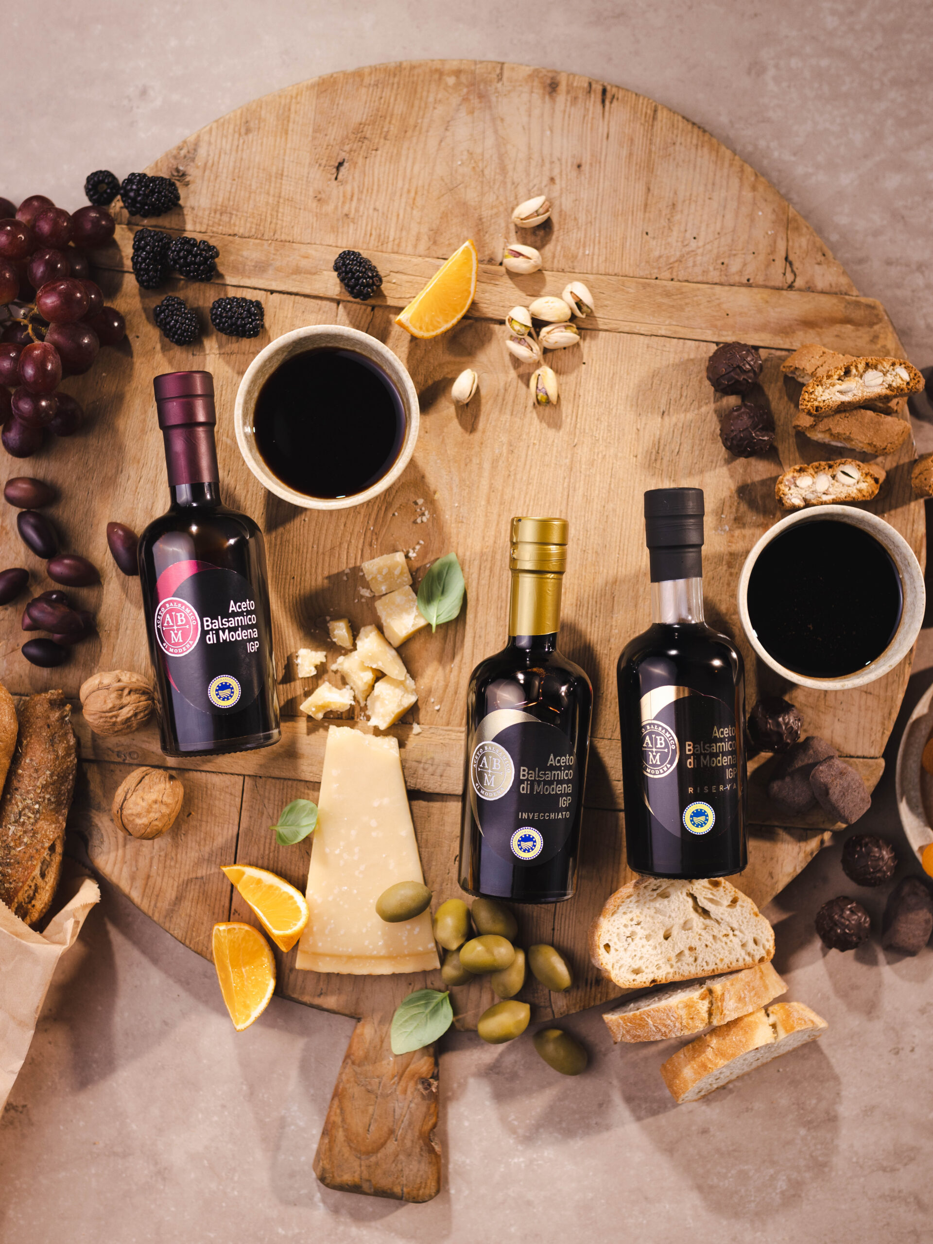 Drei Flaschen Balsamico-Essig auf einem Holzbrett, umgeben von Trauben, Nüssen und Käse.