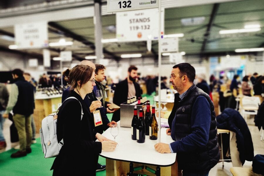 Zwei Personen im Gespräch an einem Tisch mit Weinflaschen auf einer Messe für biologische Weine.