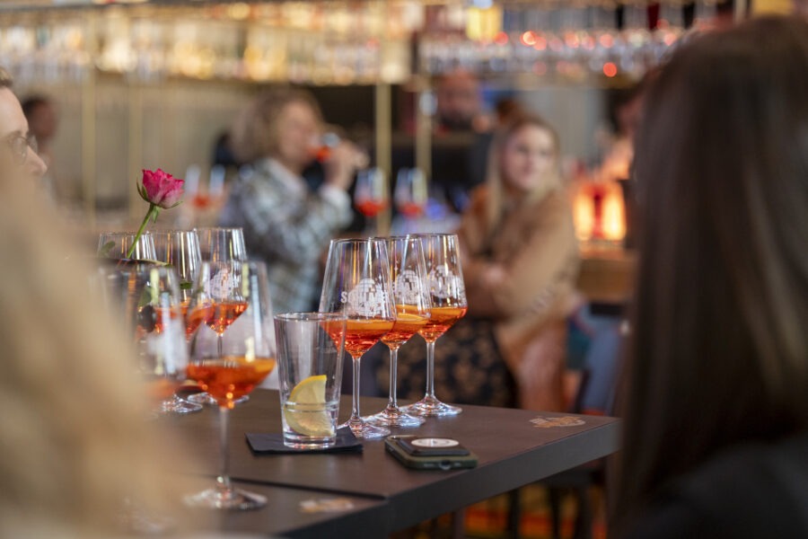 Rosé-Weingläser stehen auf einer Bar-Tafel, daneben Zitronenscheiben; im Hintergrund Gesprächende Gäste und eine rosa Blüte bei Prowein