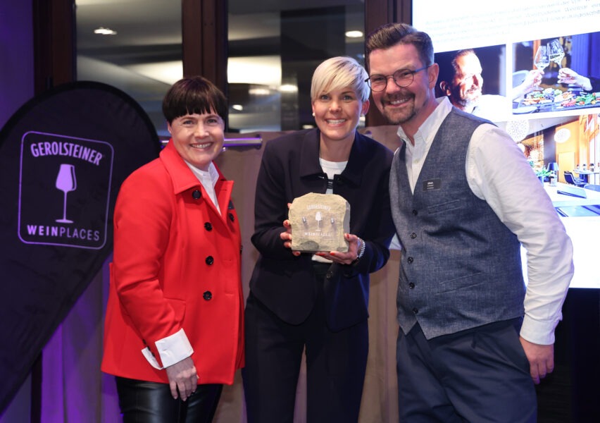 Bei der Weinpräsentation in Herford halten zwei Personen ein Gerolsteiner Weinplaces-Award; rot, schwarz und blau gekleidet, vor leuchtendem Hintergrund feierlich.