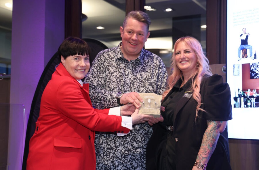 Gerolsteiner WeinPlace mit Tüte vor dunklem Hintergrund: Drei Personen posieren lächelnd, orange-roter Mantel und schwarz-weiß gemusterte Kleidung im Fokus.