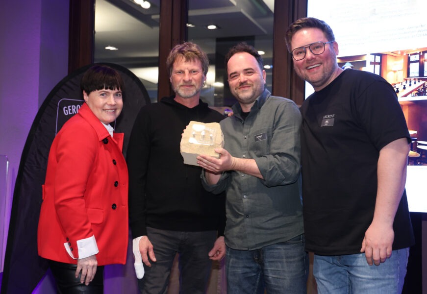 Eine Gruppe beim Gerolsteiner Wein-Place in Mainz: vier Personen, rote Jacke, Brille und dunkle Outfits, einer hält eine Weinverpackung.