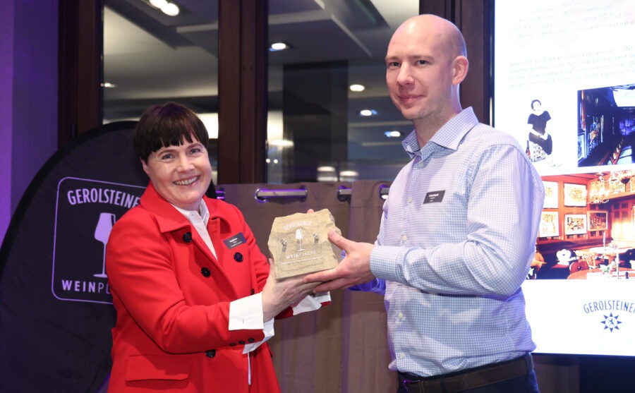 Gerolsteiner Weinplace: Zwei Personen bei der Übergabe, eine Person im roten Jackett und eine im blau karierten Hemd, im Messeumfeld.