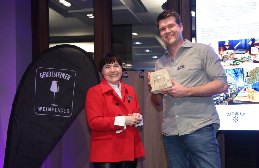 Zwei Personen mit Gerolsteiner-Weinplaces-Aufsteller und Tasche, die ein Paket mit „Weinplaces“ präsentieren; roter Mantel, schwarzer Beutel, lila Beleuchtung.