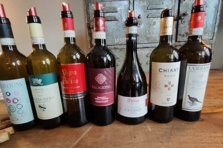 Mehrere Chianti-Weine und andere toskanische Rotweine stehen nebeneinander auf einem Holztisch vor alten Metallspinden in einem rustikalen Raum.